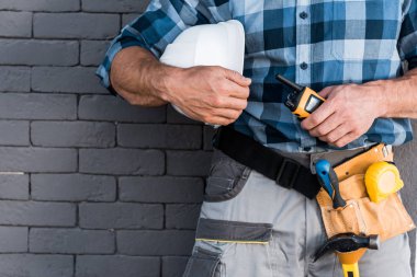 cropped view of builder holding walkie talkie and helmet near brick wall  clipart