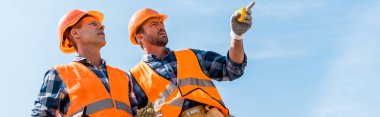 panoramic shot of handsome constructor pointing with finger while standing against blue sky  clipart