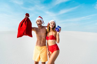 sexy and smiling girlfriend holding gift and boyfriend with santa sack on beach in Maldives  clipart