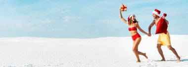 panoramic shot of sexy and smiling girlfriend holding gift and boyfriend with santa sack on beach in Maldives  clipart