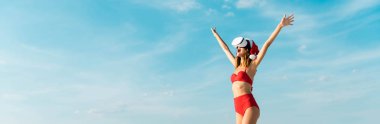 Noel Baba şapkalı ve bikinili seksi bir kadının Maldivler sahilinde sanal gerçeklik kulaklığı taktığı panoramik bir fotoğraf. 