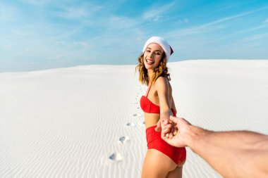 cropped view of boyfriend holding hand of sexy girlfriend in santa hat on beach in Maldives  clipart