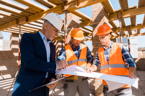 mature businessman near architects in helmets holding blueprint