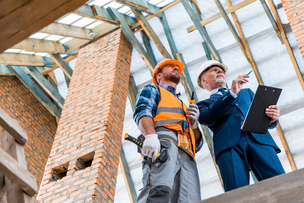 low angle view of bearded businessman holding clipboard near builder 