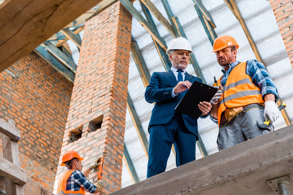 low angle view of bearded businessman holding clipboard near builders