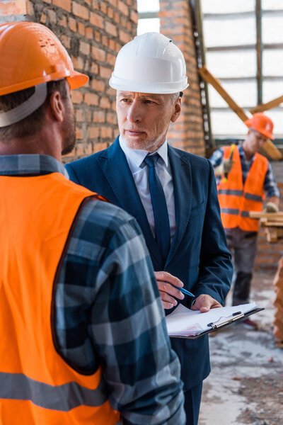 selective focus of bearded businessman in helmet holding clipboard near constructors 