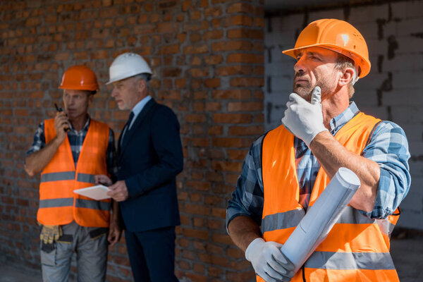 selective focus of builder holding blueprint near coworker and businessman 