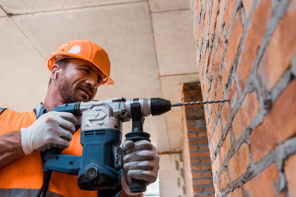 handsome man in earphones holding hammer drill near brick wall