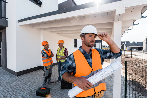 selective focus of constructor with blueprint touching helmet near men 