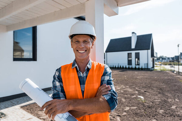cheerful mature man holding blueprint and smiling near houses 
