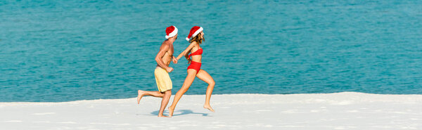 panoramic shot of sexy girlfriend and boyfriend in santa hats running on beach in Maldives 