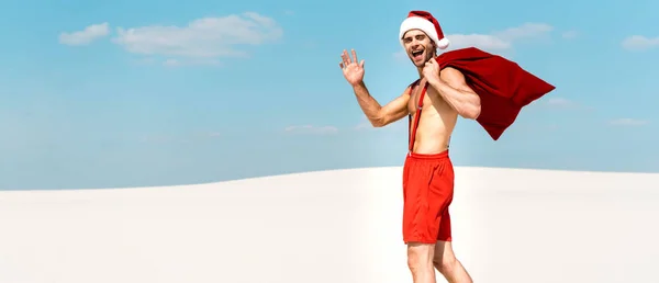 サンタの帽子のハンサムなセクシーな男のパノラマのショットサンタの袋を保持し モルディブのビーチで手を振る — ストック写真
