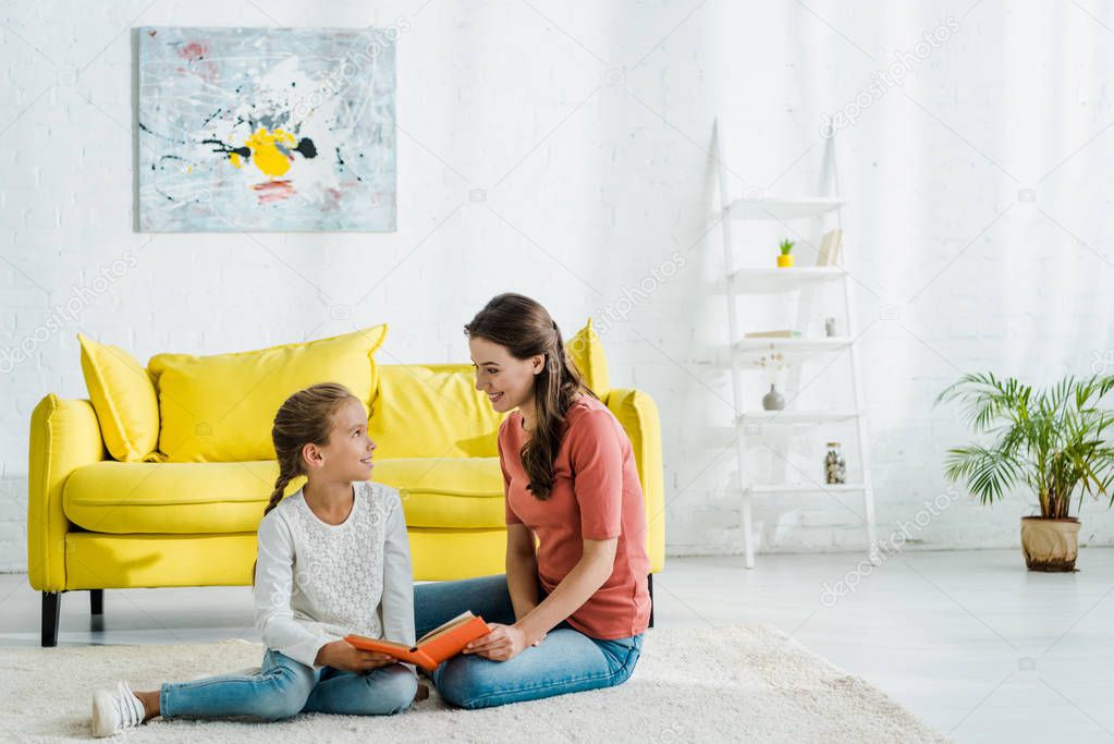 happy kid holding book while sitting on carpet with babysitter 