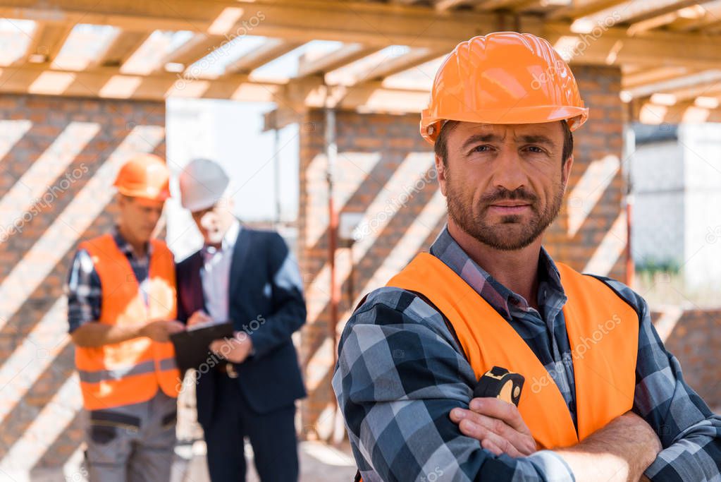 selective focus of handsome man in helmet standing with crossed arms near coworker and businessman 