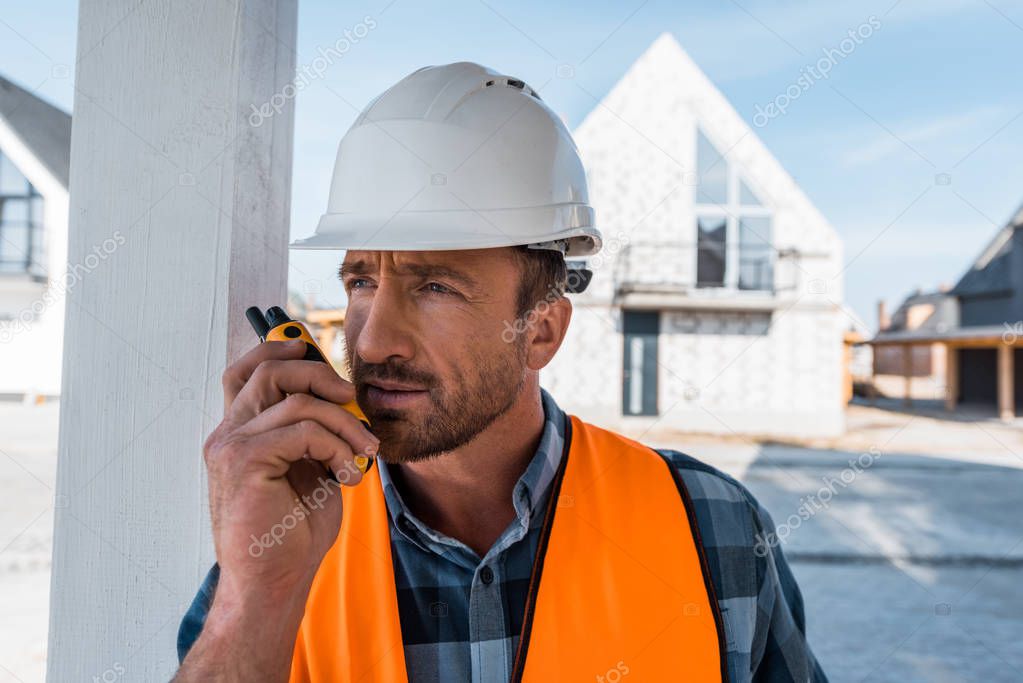 handsome bearded man in helmet holding walkie talkie near houses 