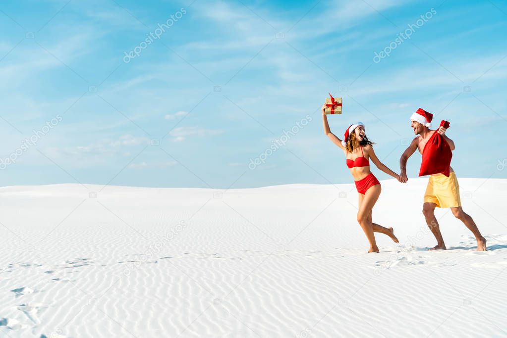 sexy and smiling girlfriend holding gift and boyfriend with santa sack on beach in Maldives 