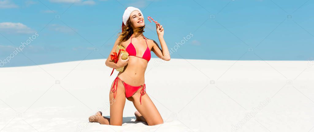 panoramic shot of sexy and smiling woman in santa hat and swimsuit holding christmas gift and candy cane on beach in Maldives 