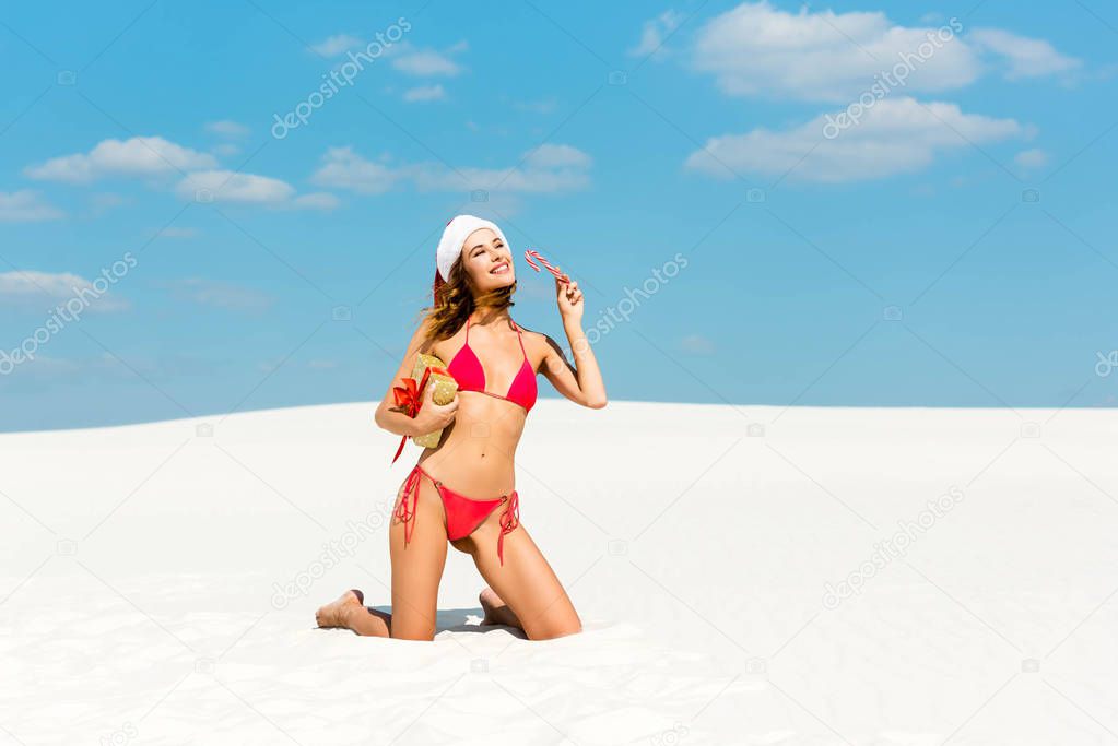 sexy and smiling woman in santa hat and swimsuit holding christmas gift and candy cane on beach in Maldives 