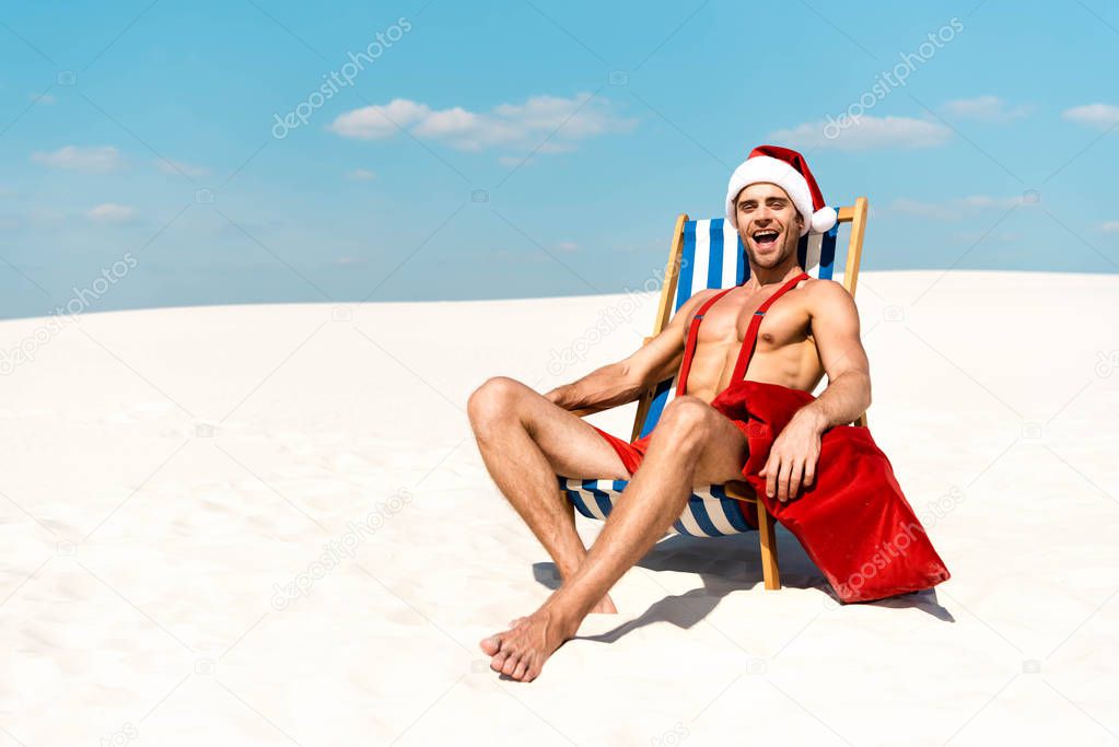 handsome and sexy man in santa hat with santa sack and sitting on deck chair on beach in Maldives 