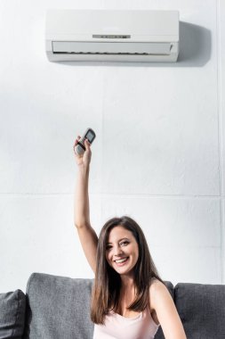 attractive and smiling woman switching on air conditioner with remote controller clipart