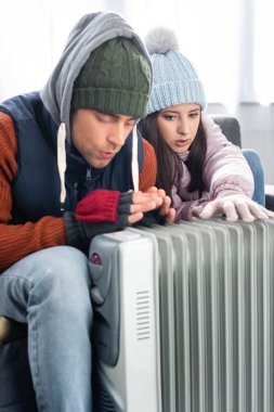 attractive girlfriend and boyfriend in winter outfit warming up near heater  clipart