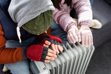 high angle view of girlfriend and boyfriend in winter outfit warming up near heater  clipart