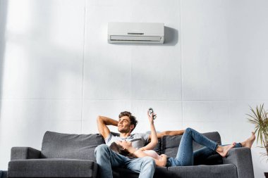 attractive girlfriend switching on air conditioner and lying on legs of handsome boyfriend  clipart