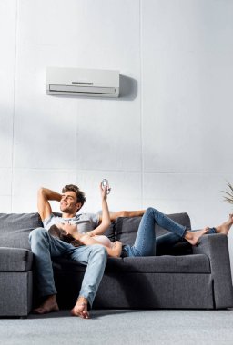 attractive girlfriend switching on air conditioner and lying on legs of handsome boyfriend clipart