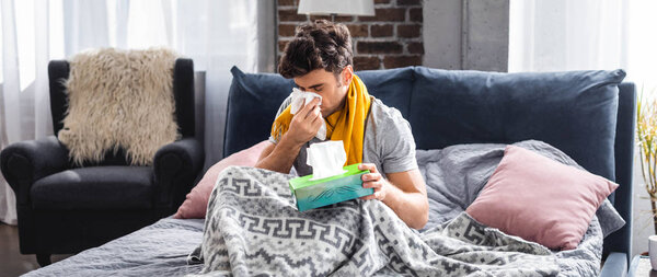 panoramic shot of sick man in scarf sneezing and holding napkin 