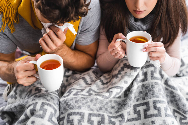 cropped view of sick girlfriend and boyfriend holding cups and sneezing with tea 