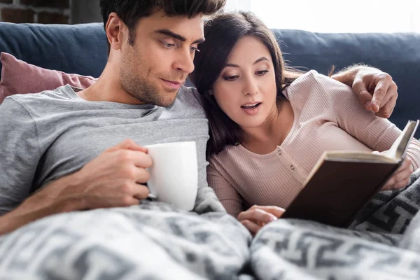 Shocked Girlfriend Reading Book Boyfriend Holding Cup Tea — Stock Photo, Image