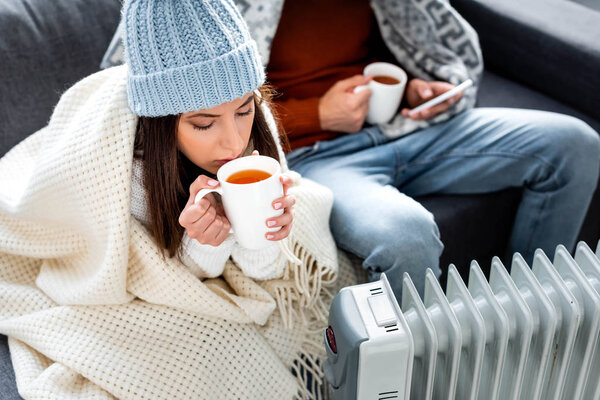 selective focus of attractive girlfriend with cup of tea warming up near heater 