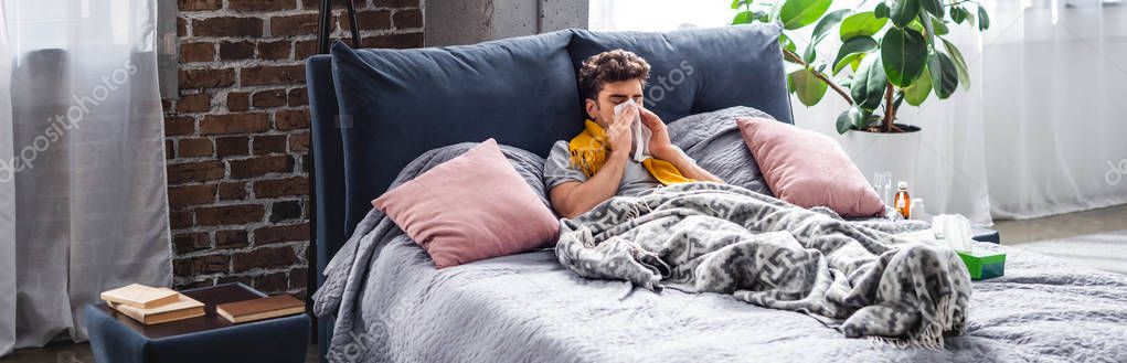 panoramic shot of sick man in scarf sneezing and lying in bed 