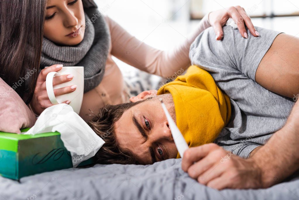 selective focus of sick girlfriend with cup and boyfriend looking at thermometer 