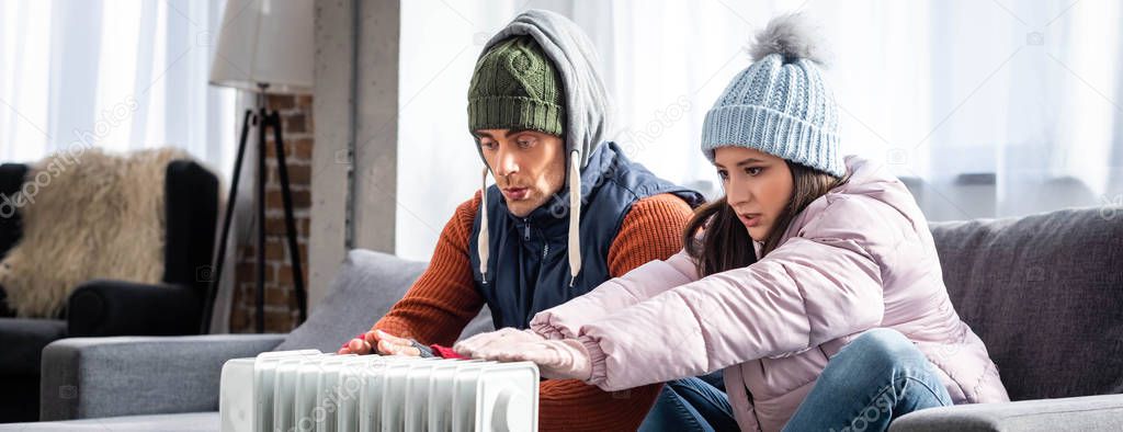 panoramic shot of girlfriend and boyfriend in winter outfit warming up near heater 