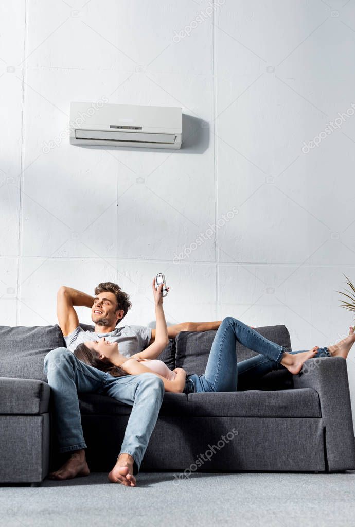 attractive girlfriend switching on air conditioner and lying on legs of handsome boyfriend