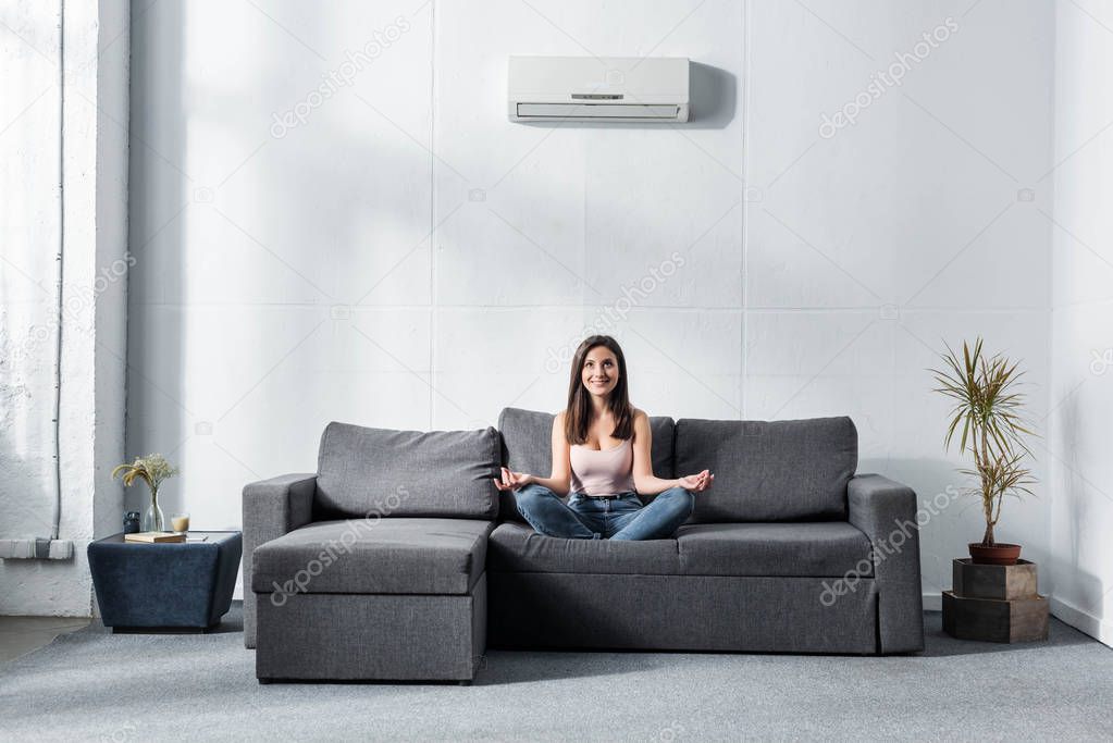 attractive and smiling woman in lotus pose sitting on sofa 