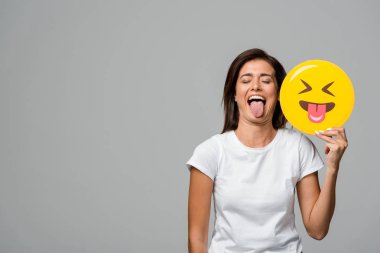 Kyiv, Ukrayna - 10 Eylül 2019: Gri renkte izole edilmiş sarı emojiyi tutarken dilini dışarı çıkaran güzel bir kadın