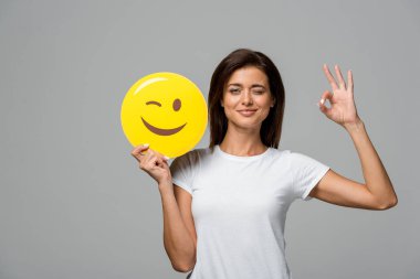 Kyiv, Ukrayna - 10 Eylül 2019: Sarı göz kırpan emoji tutan güzel bir kadın ve tamam işareti var, gri üzerinde izole edilmiş.