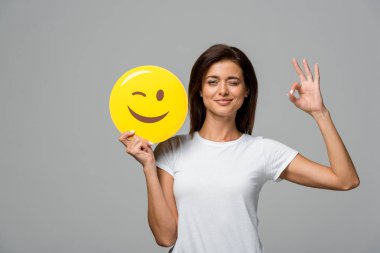 KYIV, UKRAINE - SEPTEMBER 10, 2019: beautiful woman holding yellow winking emoji and showing ok sign, isolated on grey clipart