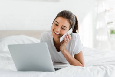 Akıllı telefondan konuşan ve sabah yatağında dizüstü bilgisayar kullanan çekici bir kız. 