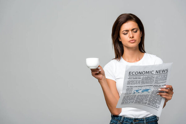 beautiful confused woman holding coffee cup while reading economic newspaper, isolated on grey