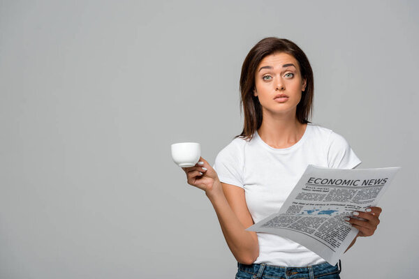 surprised young woman with coffee holding economic newspaper, isolated on grey