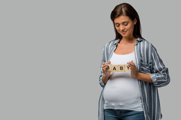beautiful happy pregnant woman holding alphabet blocks with baby sign, isolated on grey