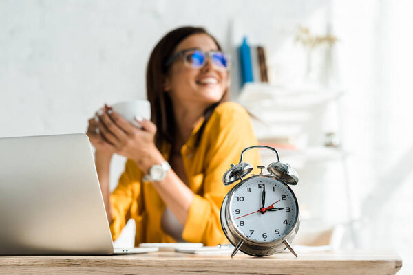 selective focus of alarm clock and happy freelancer having coffee break in home office with laptop