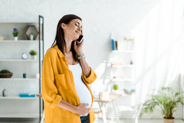 happy pregnant freelancer in yellow shirt talking on smartphone in home office