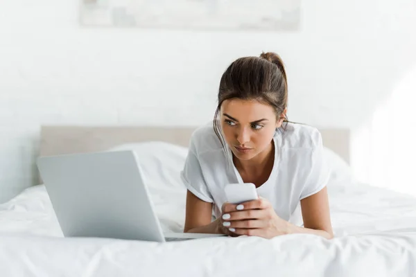 Όμορφη Σοβαρή Κοπέλα Χρησιμοποιώντας Smartphone Και Laptop Στο Κρεβάτι Πρωί — Φωτογραφία Αρχείου