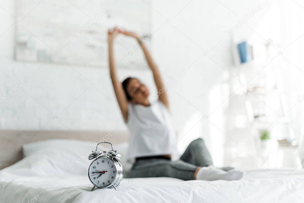 selective focus of woman stretching with alarm clock on bed in the morning