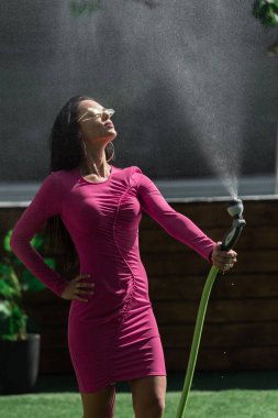 Elbiseli ve güneş gözlüklü çekici bir kadın su hortumuyla dışarıda poz veriyor. 