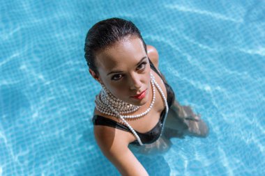 Siyah mayo ve havuzda poz veren inci kolyeli çekici bir kadının yüksek açılı görüntüsü. 